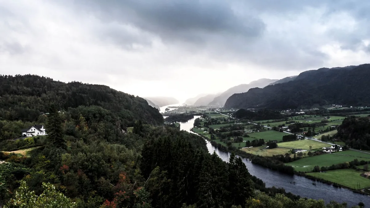 Dramatic view of a serene river valley under overcast skies, surrounded by lush green hills.